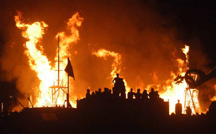 The sky is colored a bright orange as massive flames leap out while people watch.