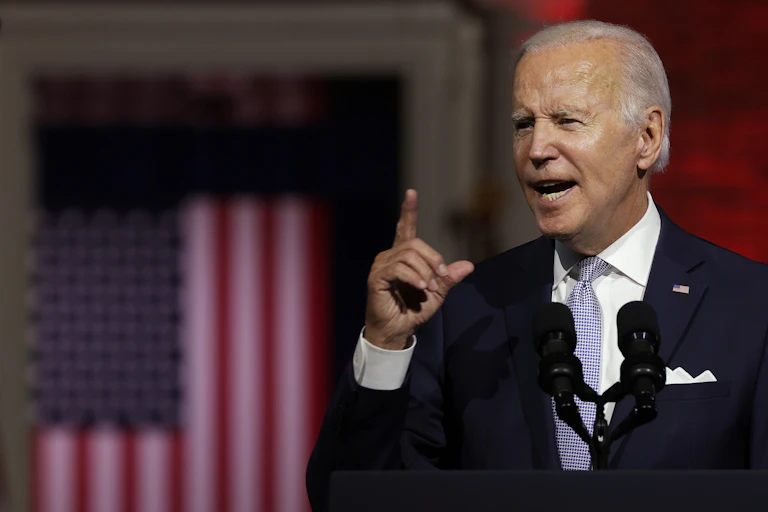 A middle-aged man dressed in a business suit raises his finger to make a point while delivering a speech in front of an American flag.