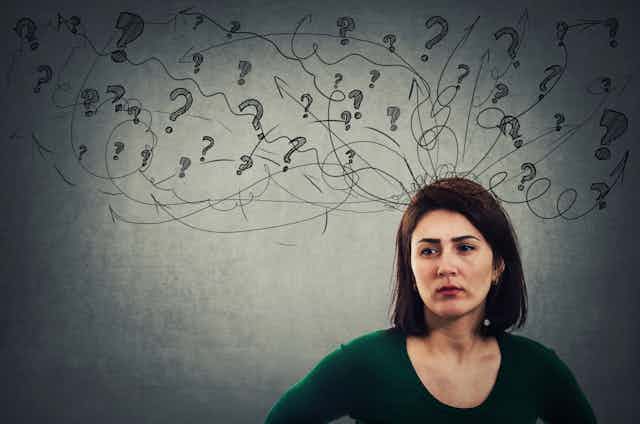 a woman stands against a grey background with a cloud of illustrated question marks above her head