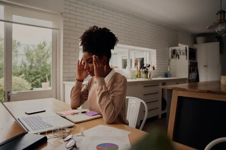 a woman sits at a kitchen table with a laptop, massaging her temples