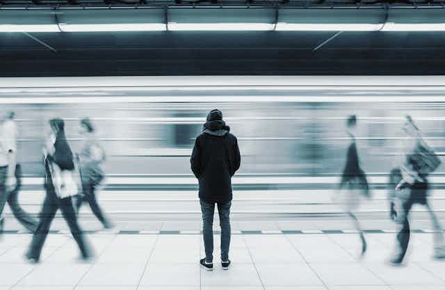 Man standing surrounded by blurry image if moving train and people walking by.