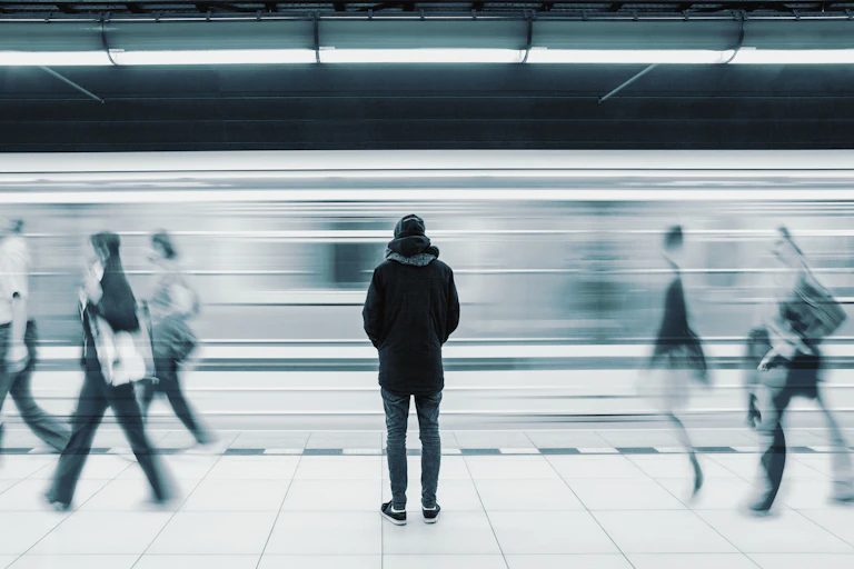 Man standing surrounded by blurry image if moving train and people walking by.