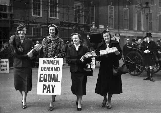 An archival photograph of four women in dress suits, holding placards and papers.