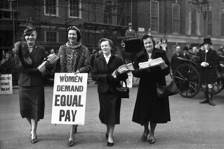 An archival photograph of four women in dress suits, holding placards and papers.