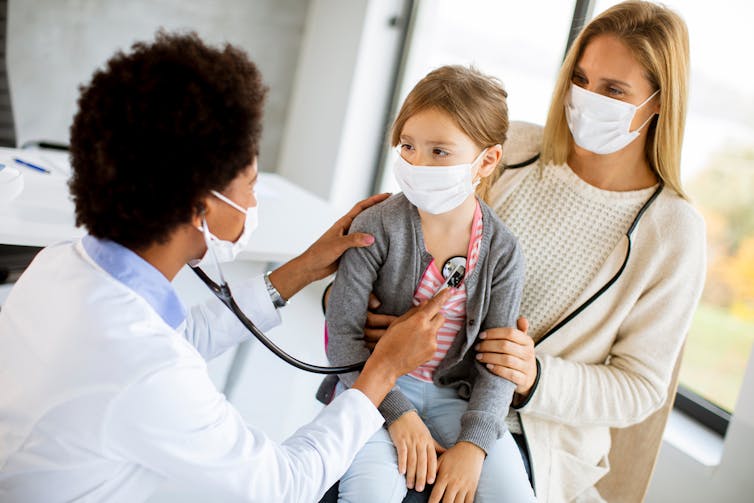 A woman in a white coat is examining a little girl using a stethoscope, while another woman stands behind the girl.