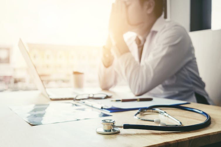 A stethoscope on a desk in the foreground, with a doctor out of focus sitting at the desk with his hands to his face