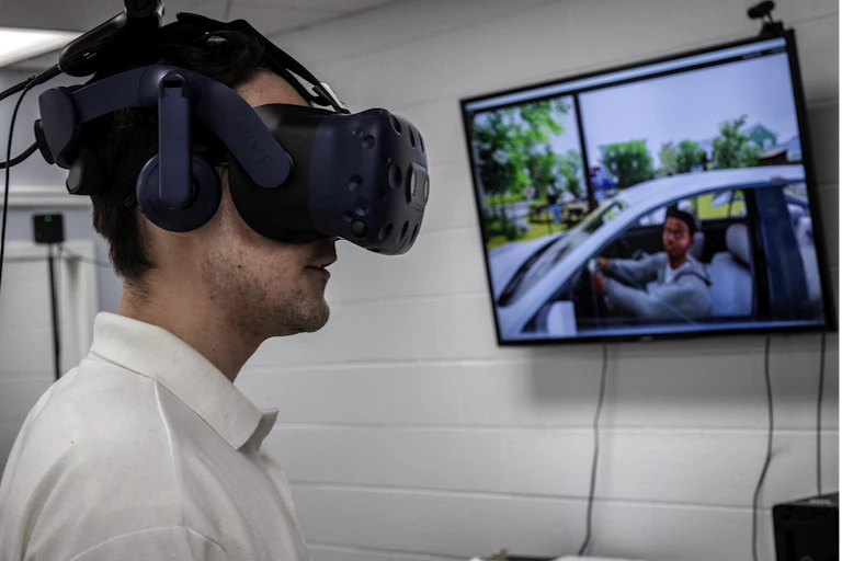A person wears a black box on his face and headphones on his ears while a screen displays what he sees.