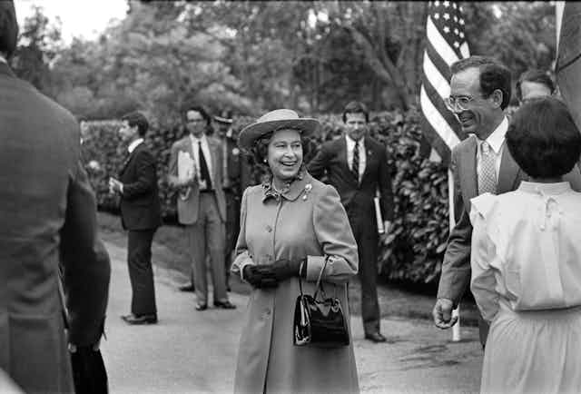 A black and white photo shows a smiling Queen Elizabeth II surrounded by men in suits and an American flag.