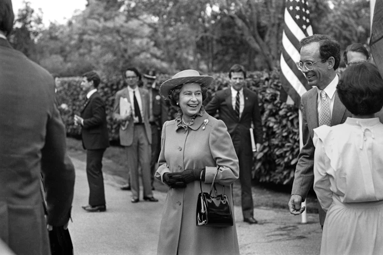 A black and white photo shows a smiling Queen Elizabeth II surrounded by men in suits and an American flag.