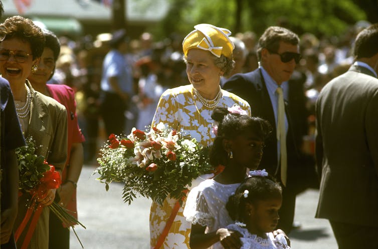 Queen Elizabeth II in a yellow and white dress and hat holds a bouquet of flowers while surrounded by well-wishers.