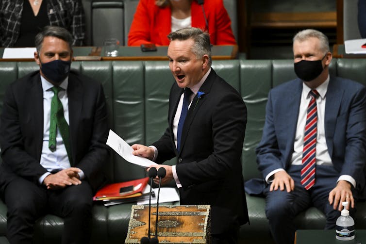 two men seated in masks watch other man speak in parliament