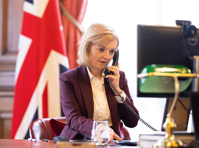 Liz Truss, the new prime minister of the UK, sitting at a desk speaking on the phone. A flag bearing the union jack is in the background.