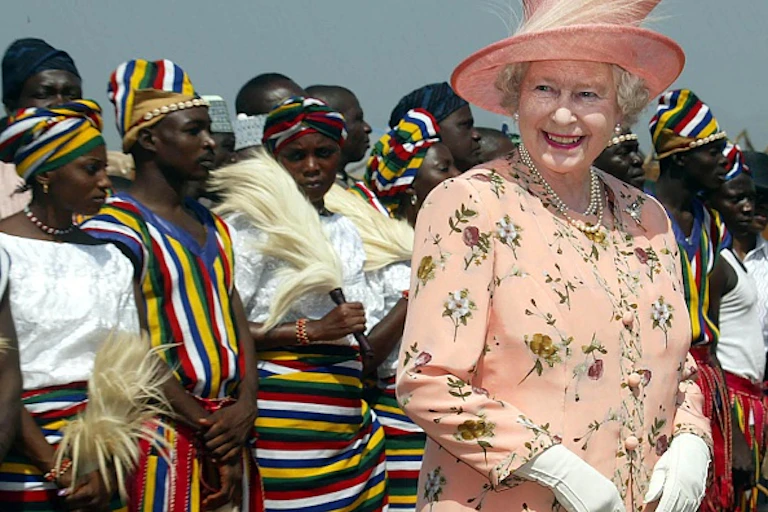 Woman in hat, pink suit and white gloves, standing and smiling in front of a goup of people in striped cloths carrying whisks