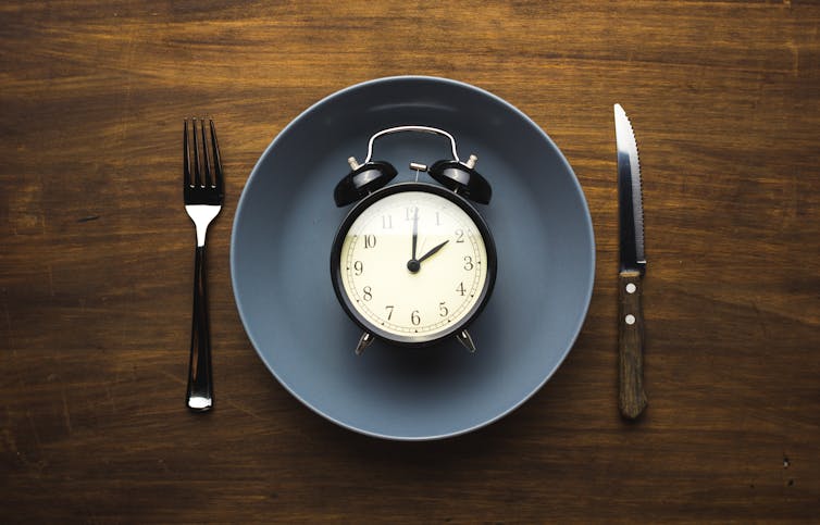 A picture of a plate on a table with a fork and knife on either side of it. In the centre of the plate is an old alarm clock.