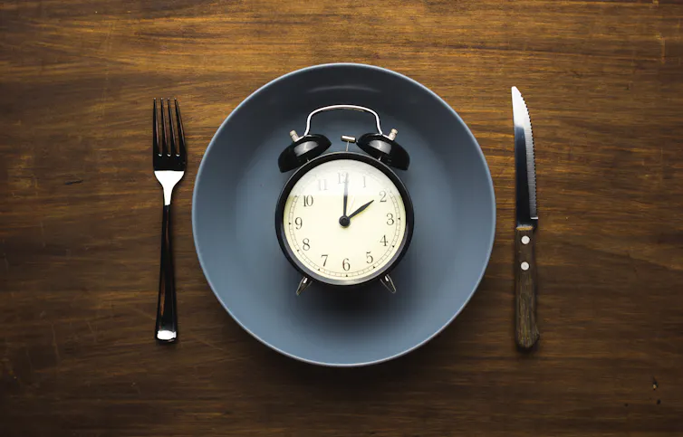 A picture of a plate on a table with a fork and knife on either side of it. In the centre of the plate is an old alarm clock.