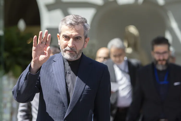 A middle aged man with salt and pepper hair and a beard wears a dark suit and waves.