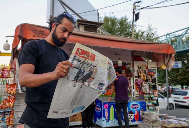 A brown skinned man with dark hair and a beard reads a newspaper in Farsi on a street, standing in front of a small store.