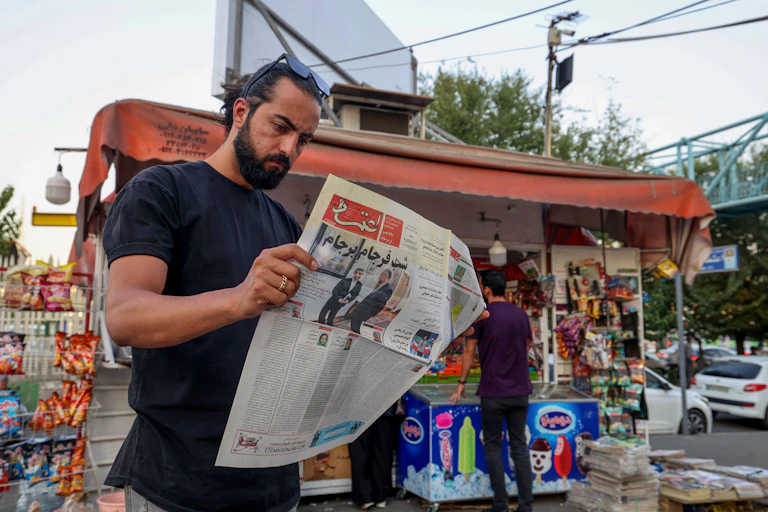 A brown skinned man with dark hair and a beard reads a newspaper in Farsi on a street, standing in front of a small store.