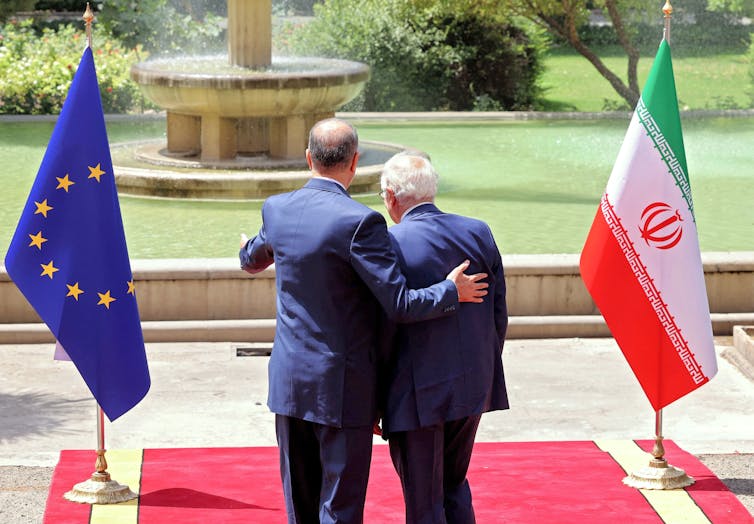 The backs of two men in suits are shown as they have their arms around each other. The European Union and Iran flags are on either side of them.