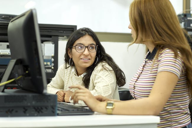 Two college age women sitting in front of a computer, one explaining something to the other.