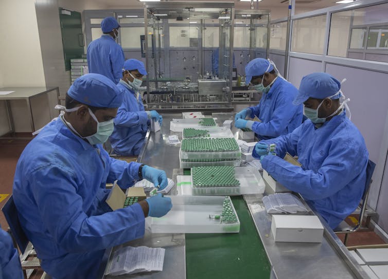 men wearing blue lab coats and face masks pack vials into boxes on a table