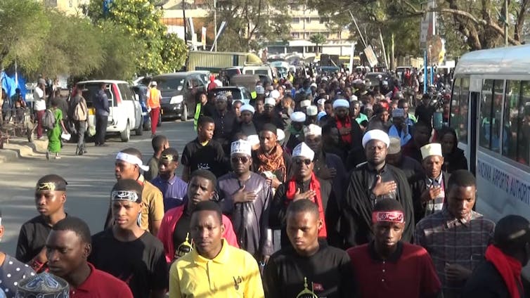 A procession on the streets of Arusha in Tanzania where most people are dressed in black with white head caps.