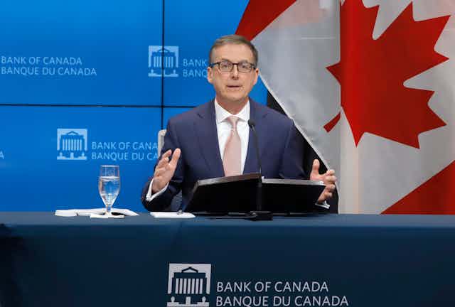 A man in a suit and glasses is seated behind a podium. He is speaking into a microphone and gesturing with his hands