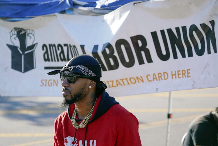 A man in a red sweatshirt stands in front of an 'Amazon Labor Union' banner.