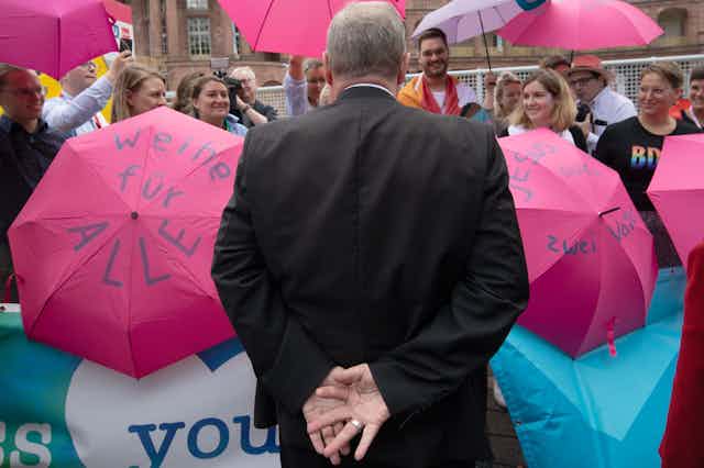 A man in a black suit with his back to the camera looks at a small crowd of people with pink umbrellas, some of which have written messages on them.