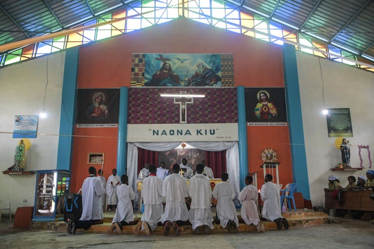 People in white robes kneel near the altar in a brightly colored church with a teal and orange wall.