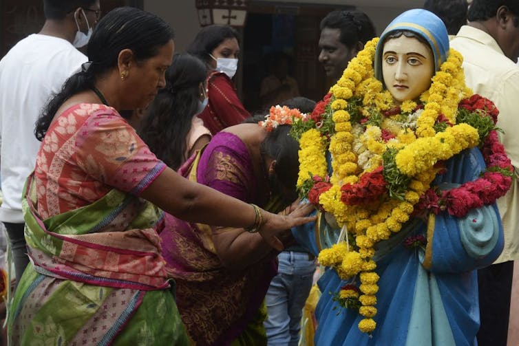 A woman in a pink shirt and green sari touches a statue of the Virgin Mary covered with garlands of flowers.