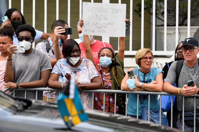 A woman in a gaggle of protesters holds a sign that reads 'Reparations Now.'