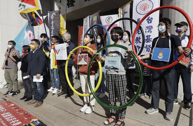 Protesters wearing masks hold protest signs and coloured hoops representing the Olympic rings.