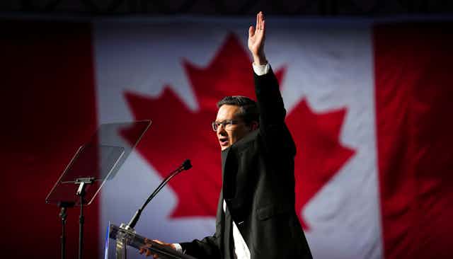 A dark haired man wearing glasses raise his arm as he speaks into a microphone. The Canadian flag is in the background.