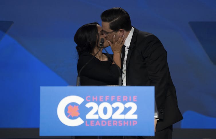 A dark-haired man and woman kiss behind a podium.