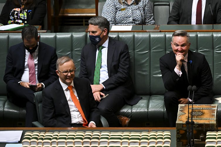 man talks in parliament as three others watch on smiling