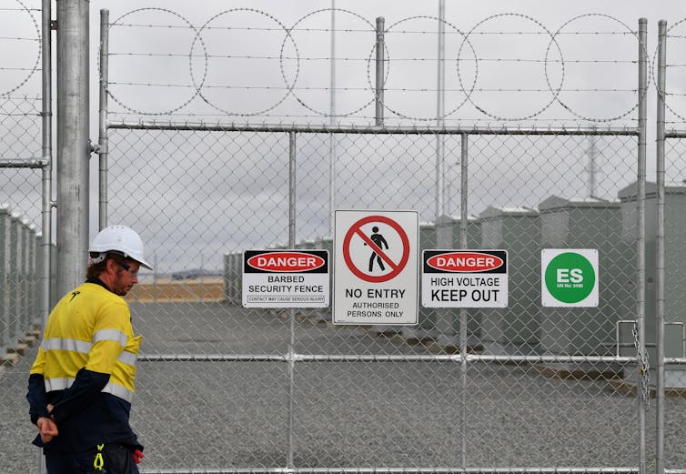 man in high-vis and hard hat stands outside fence with batteries in background