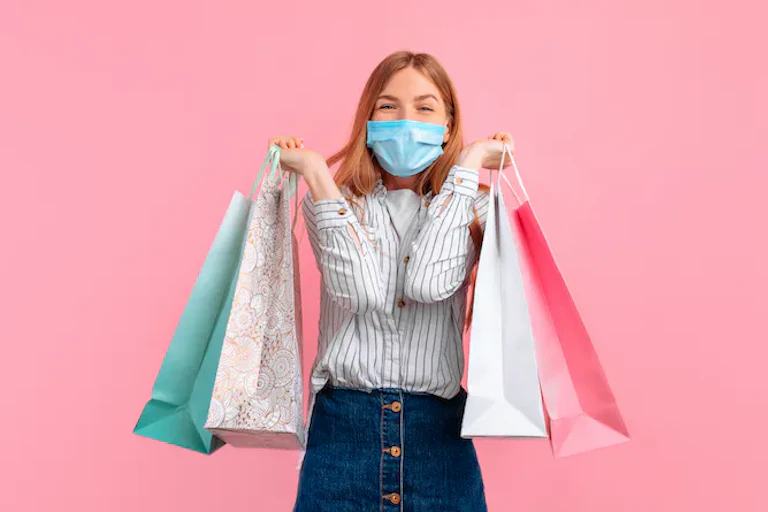 Perempuan dengan masker tersenyum dan memegang beberapa tas belanja