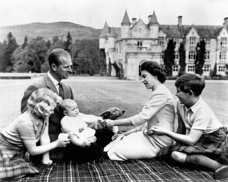 A black and white photograph of members of the royal family, seated on a tartan picnic blanket in a field.