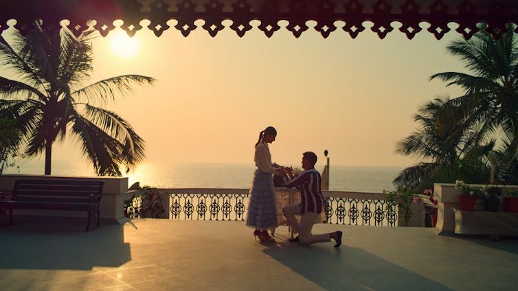 A man gets down on one knee to propose to a woman, on a beautiful balcony overlooking a sunset and the sea.