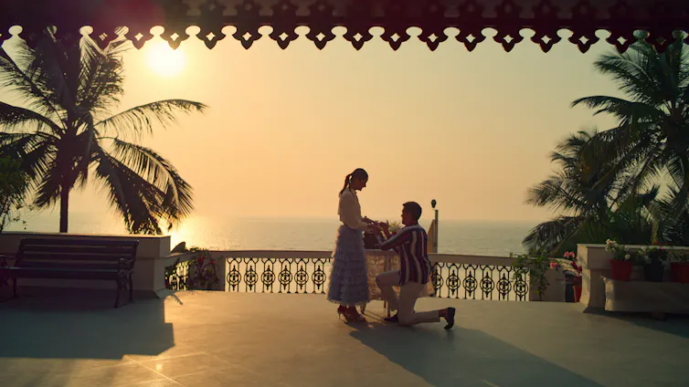 A man gets down on one knee to propose to a woman, on a beautiful balcony overlooking a sunset and the sea.