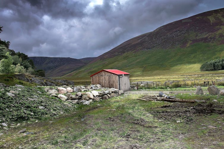 A rural hut with a red roof in mountainous countryside under a cloudy sky.
