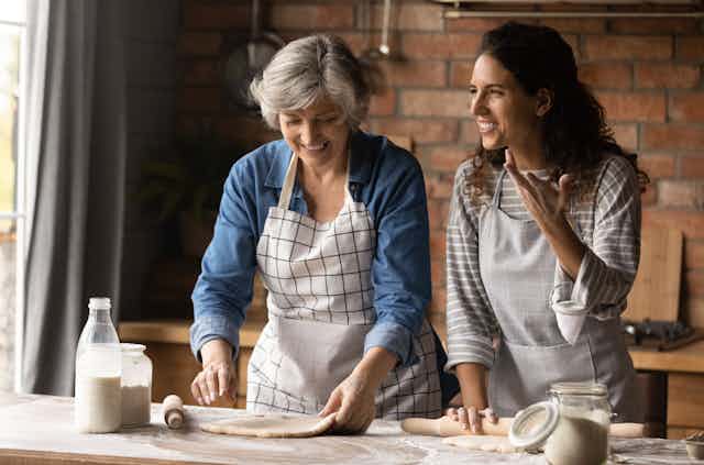 An older woman and a younger woman smiling side by side as they roll out dough for baking