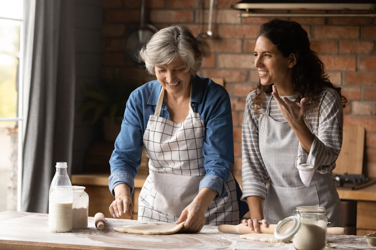 An older woman and a younger woman smiling side by side as they roll out dough for baking