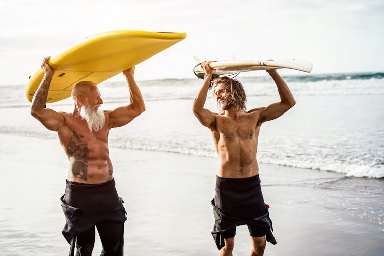 A young man and an older man holding surfboards on their heads at the beach, both wearing wetsuits and smiling at each other