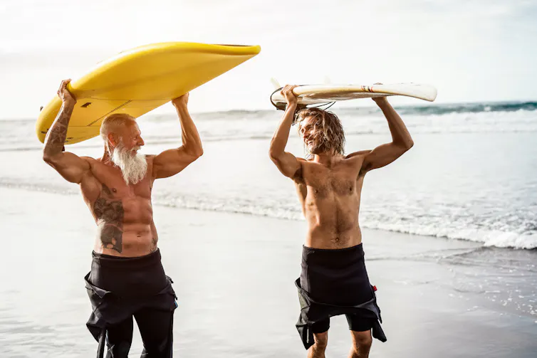 A young man and an older man holding surfboards on their heads at the beach, both wearing wetsuits and smiling at each other