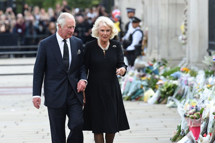الملك والملكة يرتدون ملابس سوداء ويمشون أمام صف من باقات الزهور الموضوعة.