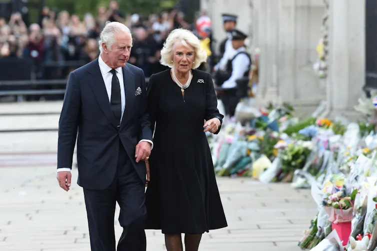 The King and queen consort dressed in black walking past a row of laid flower bouquets.