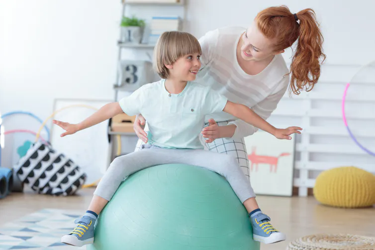 Un kinésithérapeute aide un petit garçon assis sur un ballon de gymnastique pendant une rééducation dans un hôpital pour enfants