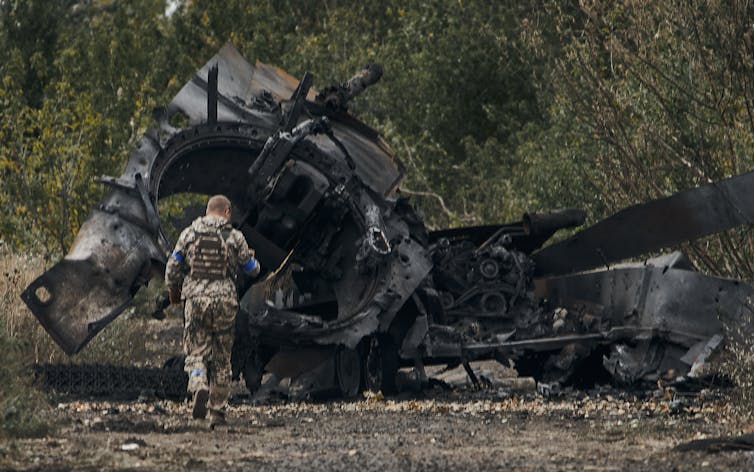 A soldier wearing camouflage garb walks in front of the burnt wreck of a tank.